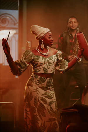 Vertical medium long shot of young Black woman wearing stylish clothes holding guiro giving jazz concert in local barの写真素材