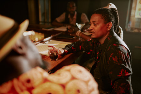 African American musician spending evening in bar after live performance, relaxing, drinking alcohol and talking to friendの写真素材