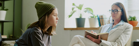 Young girl wearing beanie engaging in conversation with therapist holding notebook, seated in comfortable room with plants and sunlight filling space from windowの写真素材
