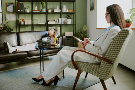 Therapist in modern office conducting session with young client lying on couch relaxation evident amidst stylish surroundings bookshelf and indoor plants visible in backgroundの写真素材