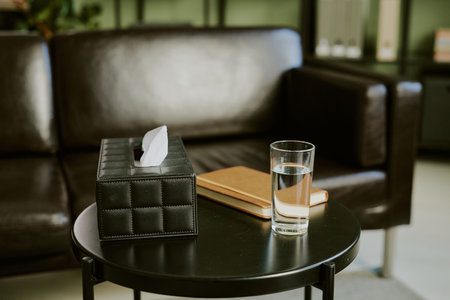 Tissue box, notebook, and glass of water placed on a round table next to a leather sofa, suggesting a cozy and organized living spaceの写真素材