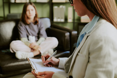 Therapist wearing suit taking notes while counseling young woman sitting on couch in office providing professional support and advice for clients mental healthの写真素材