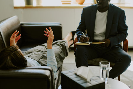 Therapist writing notes while patient lies on couch during counseling session in well-lit office with modern interior providing mental health supportの写真素材