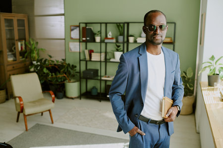 Asian man standing confidently in modern office environment holding books. Featuring well-organized office decor with bookshelves, armchair, and desk plantの写真素材