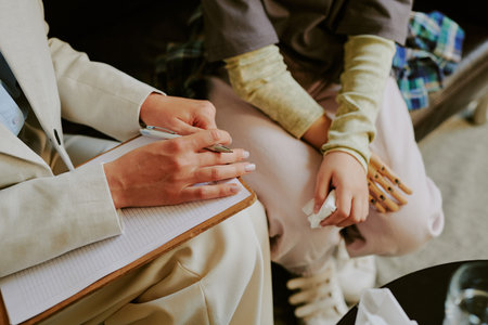 Hands of two individuals holding during therapy session, providing comfort and support to each other. Visible details include clipboard, notes, and crossed legsの写真素材