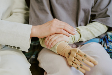Close-up of two individuals holding hands, offering comfort and support. One person has prosthetic limb, showcasing moment of connection and careの写真素材