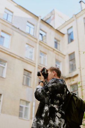 Male photographer standing outdoors and holding camera while taking picture of urban scene. Backdrop consists of tall, light-colored buildings in city environmentの写真素材