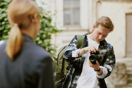 Young man holding camera and making adjustments while standing in outdoor setting with greenery in background and another person nearby focusing on himの写真素材