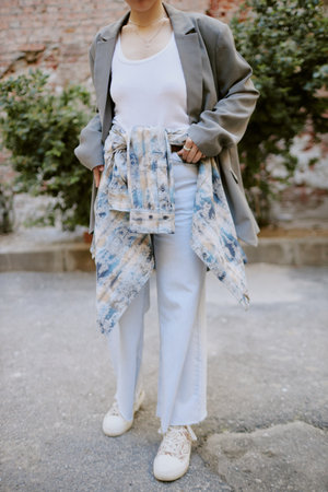Individual standing in outdoor setting, wearing a grey blazer over a white tank top, paired with white pants, sneakers, and a tie-dye shirt tied around waistの写真素材