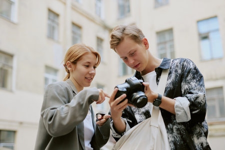 Young individuals examining camera settings in outdoor cityscape, surrounded by buildings, one holding professional camera while other points at screen, showcasing interest in photographyの写真素材