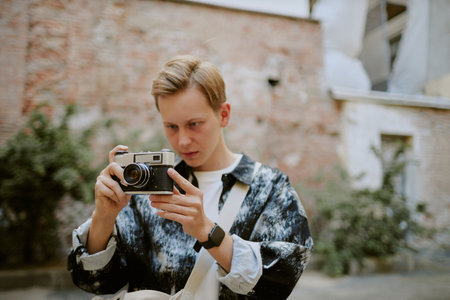 Young Caucasian man adjusting vintage camera outdoors, preparing for shot in urban environment with greenery and rustic walls behindの写真素材