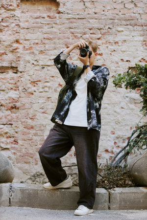 Person holding camera up to their face, standing outdoors against a brick wall, engaging in photography activity while looking up slightly in relaxed postureの写真素材