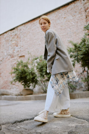 Young Caucasian woman enjoying outdoor stroll by a brick wall, dressed in casual attire and smiling, with a relaxed demeanor against an urban backgroundの写真素材