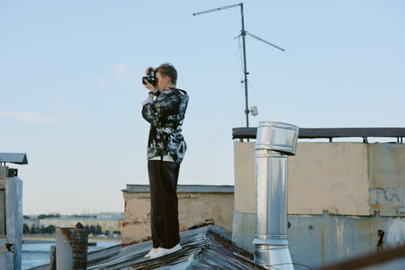 Individual standing high on rooftop and using camera to capture scenery, with antenna and chimney visible. Urban buildings in background and sky overheadの写真素材