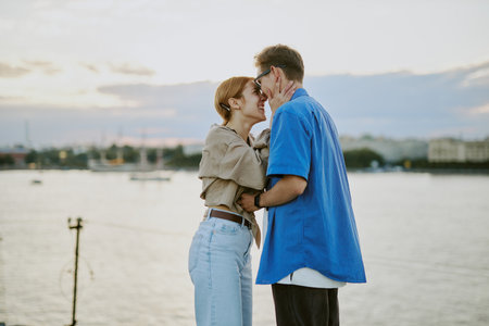 Young couple sharing intimate moment by waterfront during sunset, with city skyline in background. Casual attire and natural surroundings lending romantic feelの写真素材