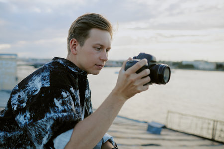 Young Caucasian man focusing camera by river with cityscape in background, wearing artistic jacket and concentrating on taking perfect shotの写真素材