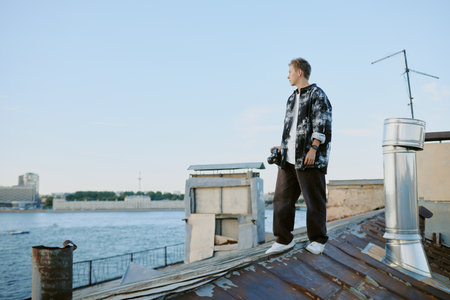 Young man standing on rooftop near waterfront, gazing into distance with thoughtful expression, holding phone in hand, showcasing modern urban lifestyle and reliance on technologyの写真素材