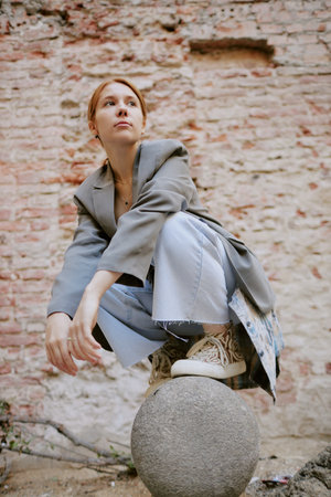 Contemplating woman in casual attire sitting on a concrete ball against a rustic brick wall background, deep in thought, with serene facial expressionの写真素材