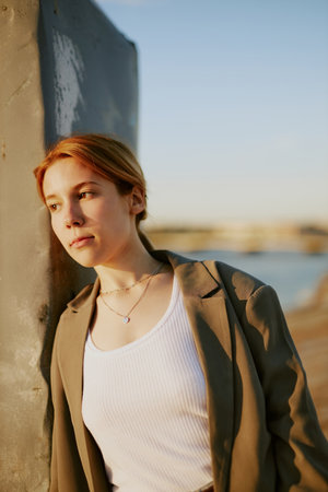 A young woman with light hair leaning against concrete column at seaside during sunset, gazing into...