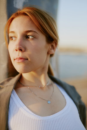 Young woman with red hair standing outside, gazing thoughtfully into distance. Wearing necklace and white top, sunlight softly illuminating her face, with blurred background adding depthの写真素材