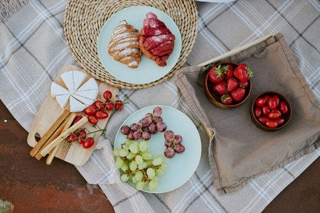 Top view of a breakfast setup featuring croissants, camembert, grapes, strawberries, and cherry tomatoes on a neutral plaid tablecloth creating a cozy and inviting atmosphereの写真素材