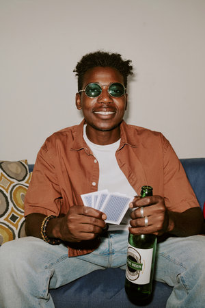 Vertical shot of young man with cards and bottle of beer sitting on sofa during card gameの写真素材