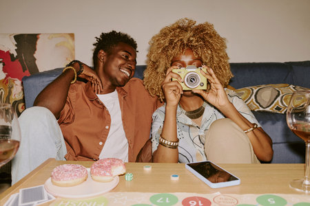 Black woman hiding her face while taking shot of her friend, black man sitting next to her and...