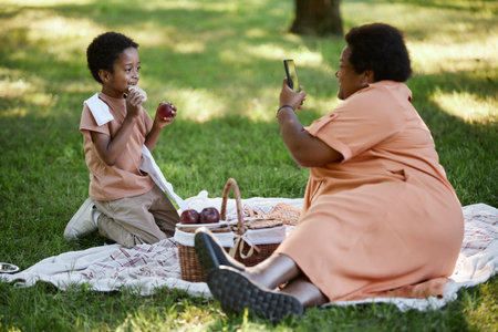 Granny taking shot of her happy grandson who eating fruits while they having picnic in green parkの写真素材
