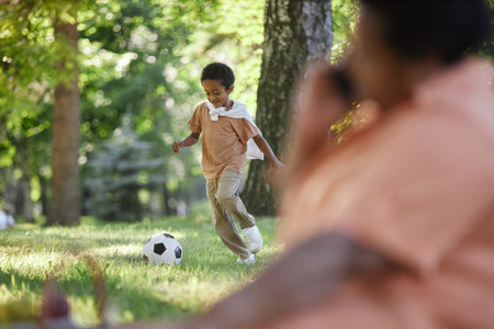 Little African American boy playing football on lawn while his grandmother speaking on phone in blurred foregroundの写真素材