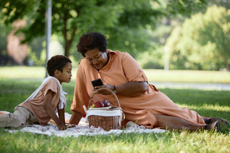 Granny and her grandson sitting on blanket and looking at phone screen while having picnic in parkの写真素材