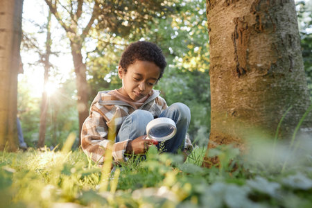 African American boy holding loupe while sitting on green grass in wood, he zooming trees rootの写真素材