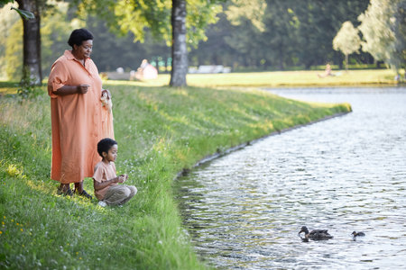 African American branny and her lovely grandson feeding ducks with white bread in green parkの写真素材