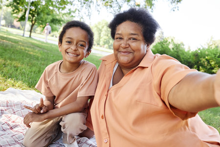 African American granny and her lovely grandson taking selfie while resting on checkered blanket in parの写真素材