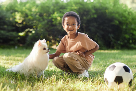 Portrait of happy African American boy caressing his snow white Pomeranian dog while walking in parkの写真素材
