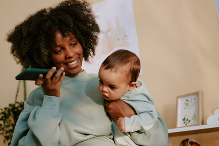 Smiling African American mom holding her baby boy while listening to voice message via phoneの写真素材