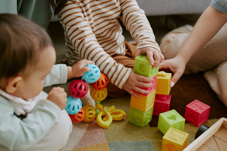 High angle shot of kids hands holding multicolored bricks and rattle while adult hand assisting them to built towerの写真素材