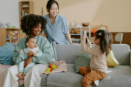 Little Girl sitting on sofa and holding photo while taking photo of her mom and her African American lifelong friend with her infant sonの写真素材