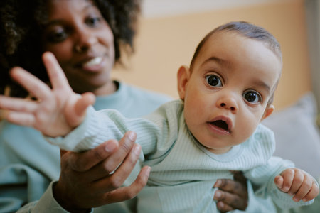 Curious baby boy looking directly at camera while trying to catch it, his African American mom...