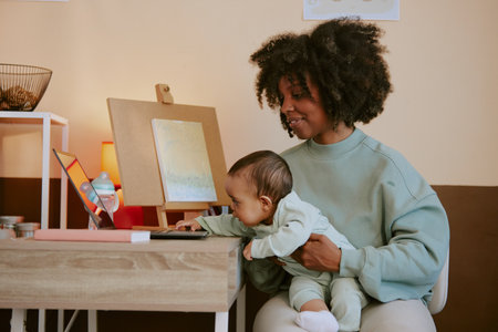 African American mother sitting with her baby while he trying to touch black laptop on tableの写真素材