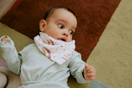 Male infant dressed in pastel blue onesie looking around nursery while lying on soft carpetの写真素材