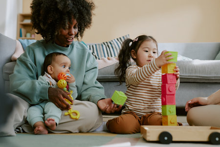 African American mom holding her baby son while helping Asian toddler to build brick towerの写真素材