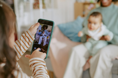 Medium close up of small hands of girl holding phone to take photo of baby with his African American mother while they sitting on sofaの写真素材