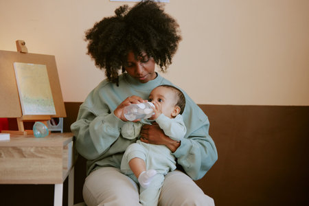 African American mother holding her adorable son and taking him baby bottle of water while he...