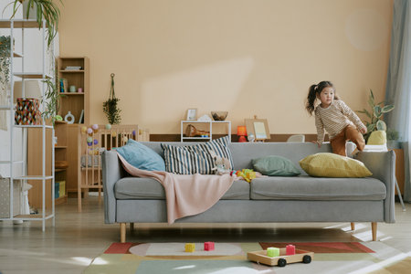 Cute little Asian girl fooling around on soft gray sofa in light beige nursery equipped with toys and furnitureの写真素材