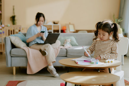 Asian little girl improving her drawing skills while her mother checking her phone in blurred backgroundの写真素材