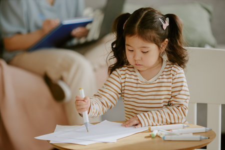 Little Asian girl sitting at wooden table and using orang felt-tip to draw picture in nursery, her mother making notions in blurred backgroundの写真素材