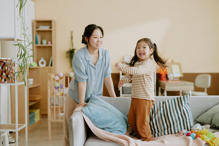 Asian little girl playing on sofa and looking directly at camera while her mother smiling and watching her in light nurseryの写真素材