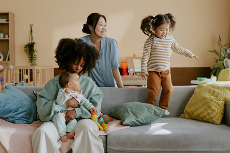 Asian mom watching her daughter jumping on sofa while her African American friend hugging her baby boy who looking at little girlの写真素材