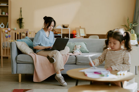 Little Asian girl sitting at wooden table and drawing while her mother working remotely on sofaの写真素材