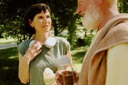 Senior brunette woman adjusting her headphones while talking to her silver-haired companion who drinking waterの写真素材